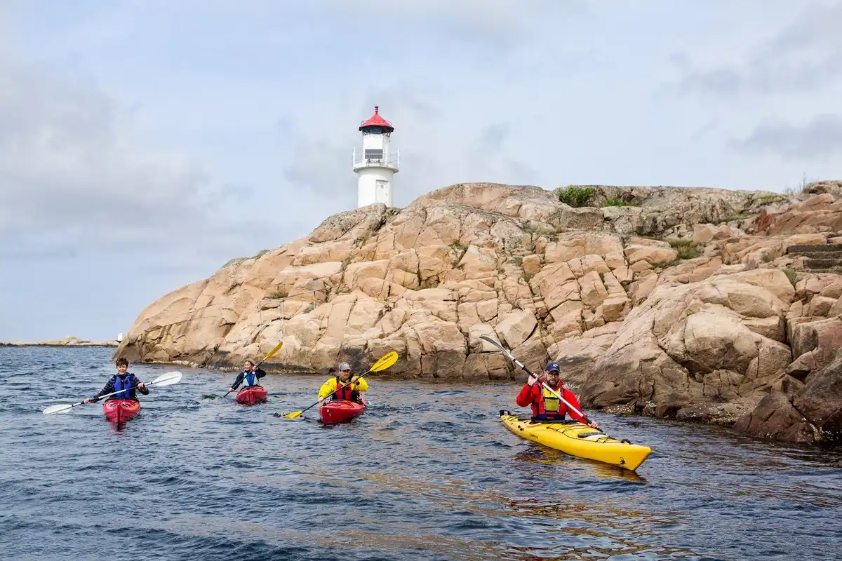 En kajakpaddlare navigerar lugnt i en havskajak nära en historisk fyr, med kustlandskapet i bakgrunden, vid Ramsvik Stugby & Camping.