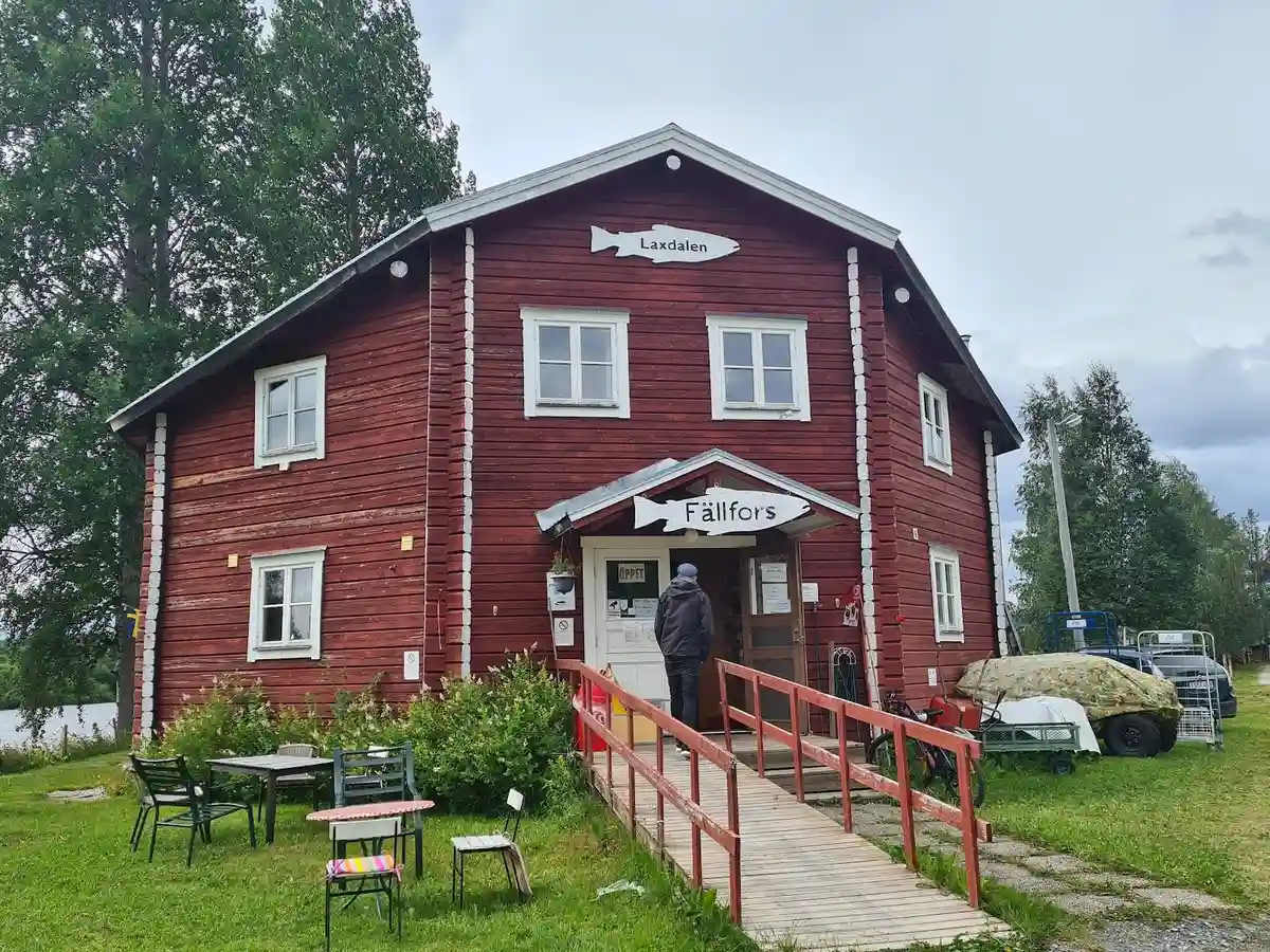 En röd stuga med vita knutar står omgiven av grönska på Fällfors Camping. Stugan har en dörr och flera spröjsade fönster som vätter ut mot en välskött gräsmatta.