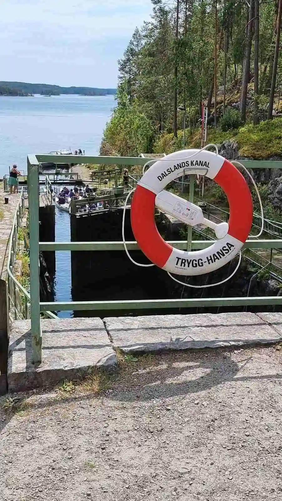 En ringboj hänger på en brygga med båtar i bakgrunden, omgiven av ett naturskönt område med sjöar och skogar vid Elovsbyn Camping & Canoe.