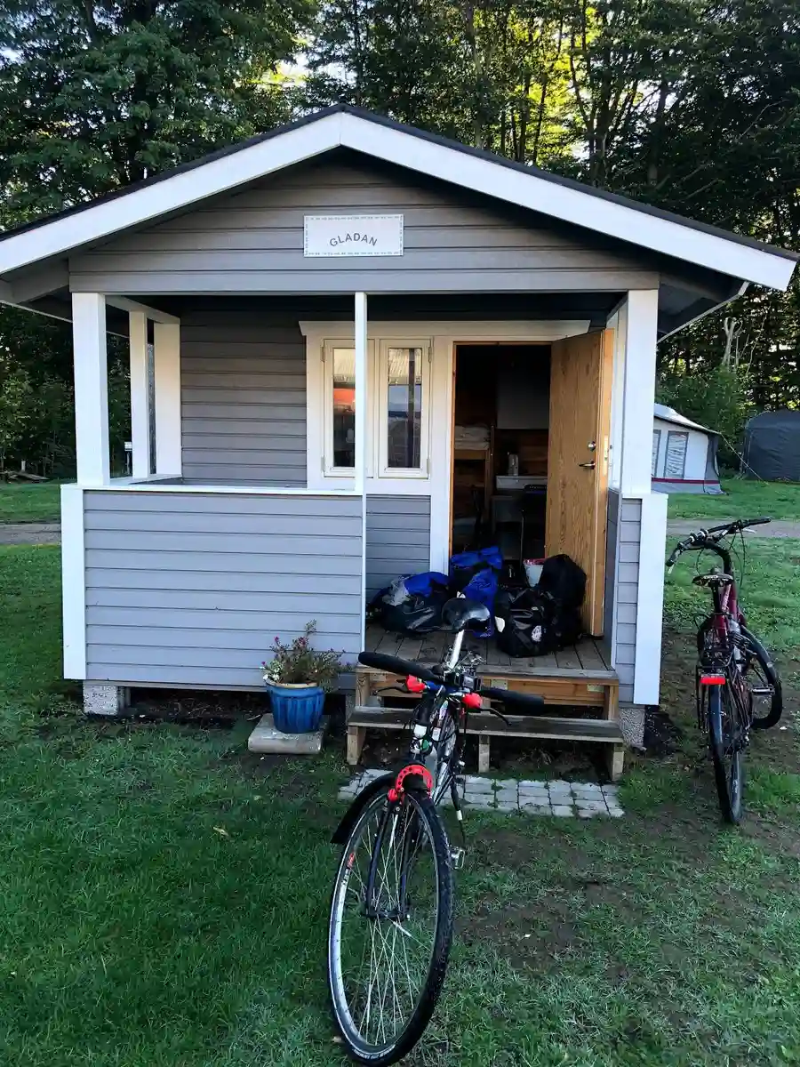 En mysig trästuga vid Elfdalens Camping, omgiven av lummiga träd och grönska, med en enkel dörr och fönster, perfekt för naturälskare och friluftsentusiaster.