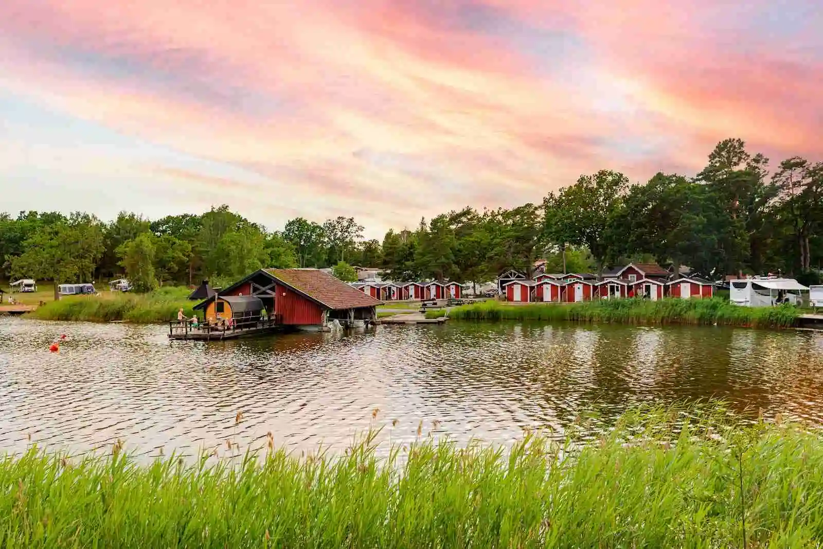 En naturskön vy över Västervik Resort med en idyllisk sjö, stugor och en båt vid strandkanten. Reflektioner i vattnet förstärker den lantliga atmosfären, omgiven av grönska och lugn.