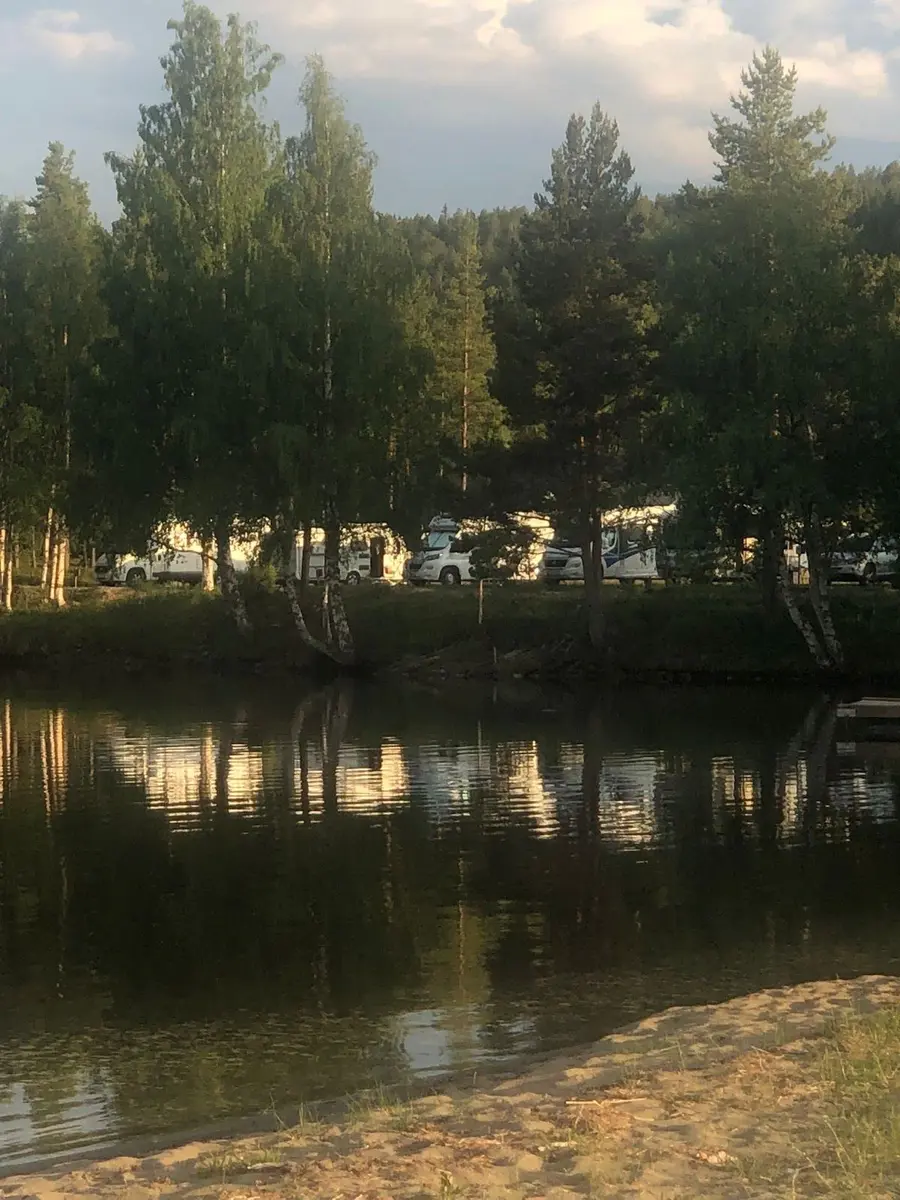 Bild på kojbyns camping: En lugn vattenyta omgiven av grönska. Vattendraget reflekterar den omgivande naturen, vilket skapar en harmonisk och fridfull atmosfär.
