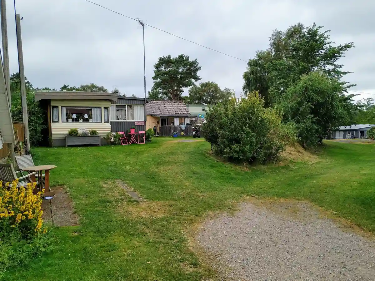 En grön campingplats med gräs och några hus, omgiven av molnig himmel och en lantlig miljö.