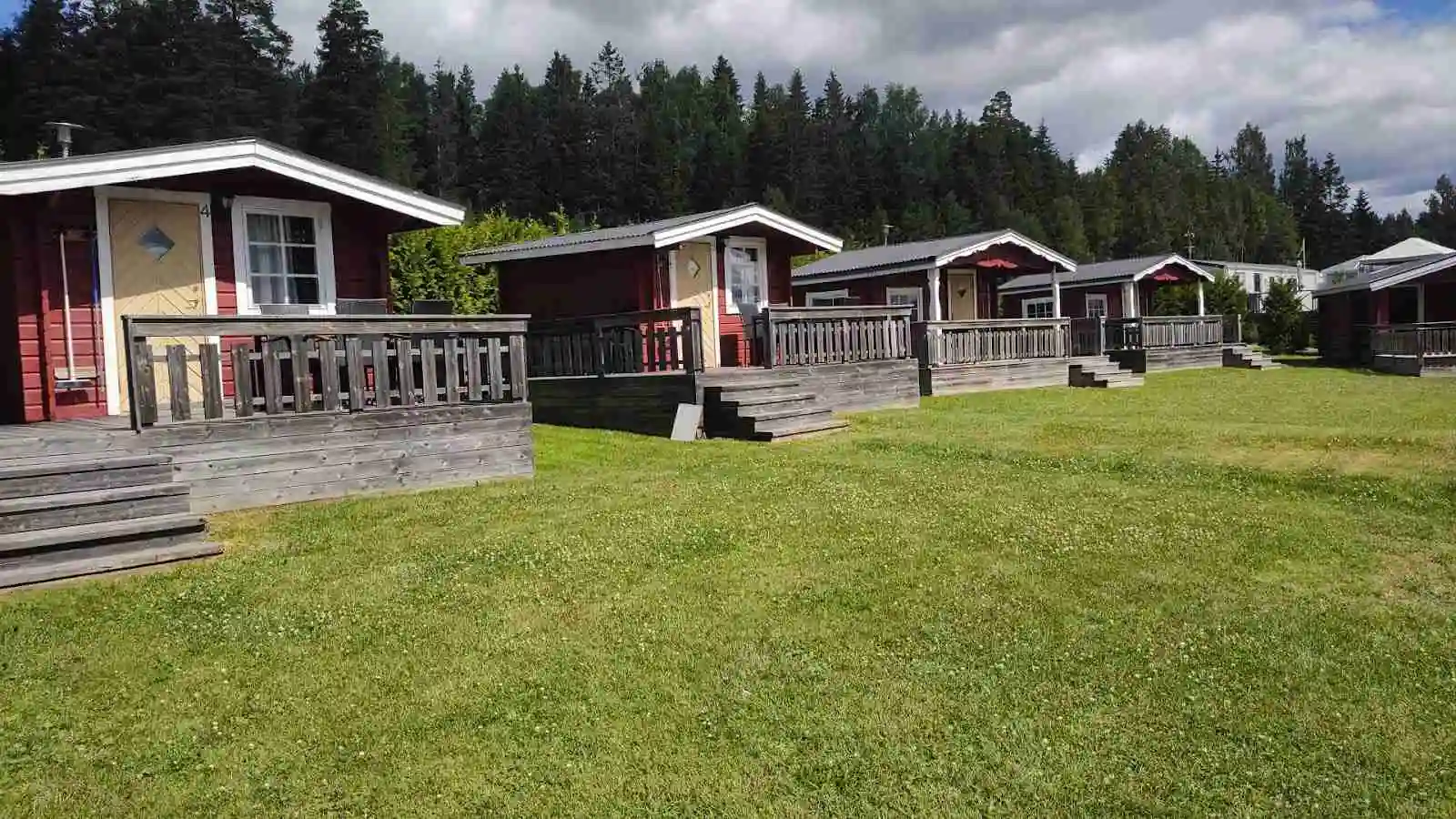 En röd stuga med veranda omgiven av en grön gräsmatta och trädgårdsmöbler i trä, belägen på Charlottenberg camping & stugor - Haganäset.