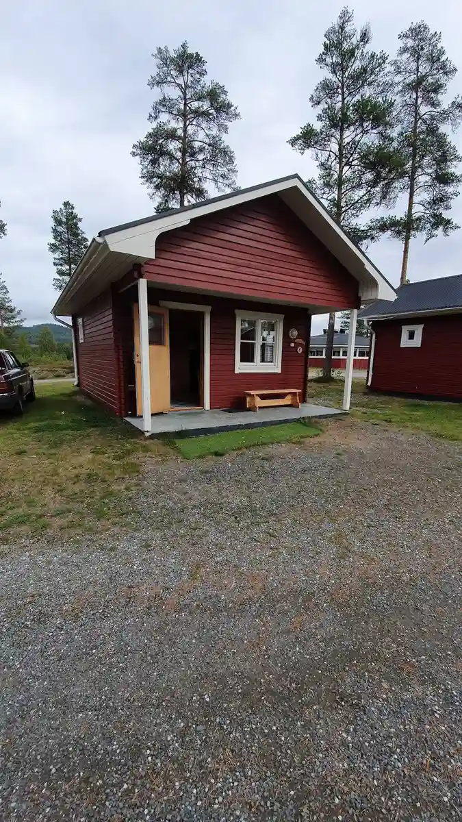 En stuga med träfasad på Fromhedens camping, omgiven av en välskött gräsmatta och inkörsel, med flera små uthus i bakgrunden.