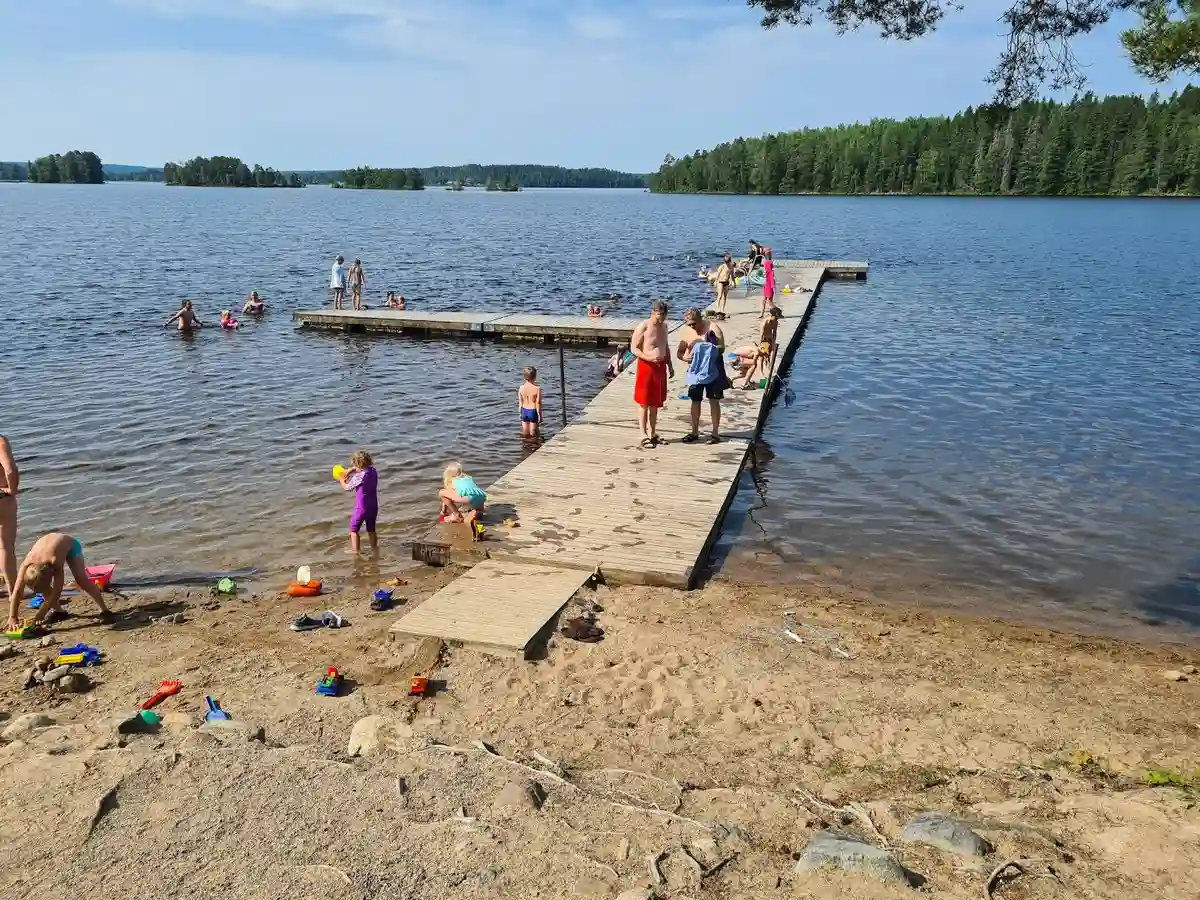 En vacker vy över Uskavigårdens camping med en sjö i förgrunden, omgiven av grönskande natur och strandlinje som inbjuder till rekreation och avkoppling.