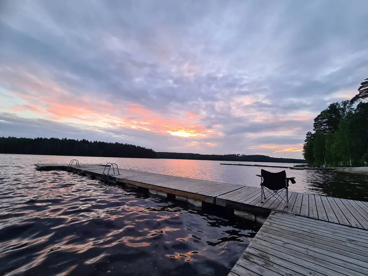 En stilla kvällsvy över Uskavigårdens camping, med en sjö i förgrunden och en brygga som sträcker sig ut i vattnet. Horisonten syns i bakgrunden under en mjukt färgad kvällshimmel.