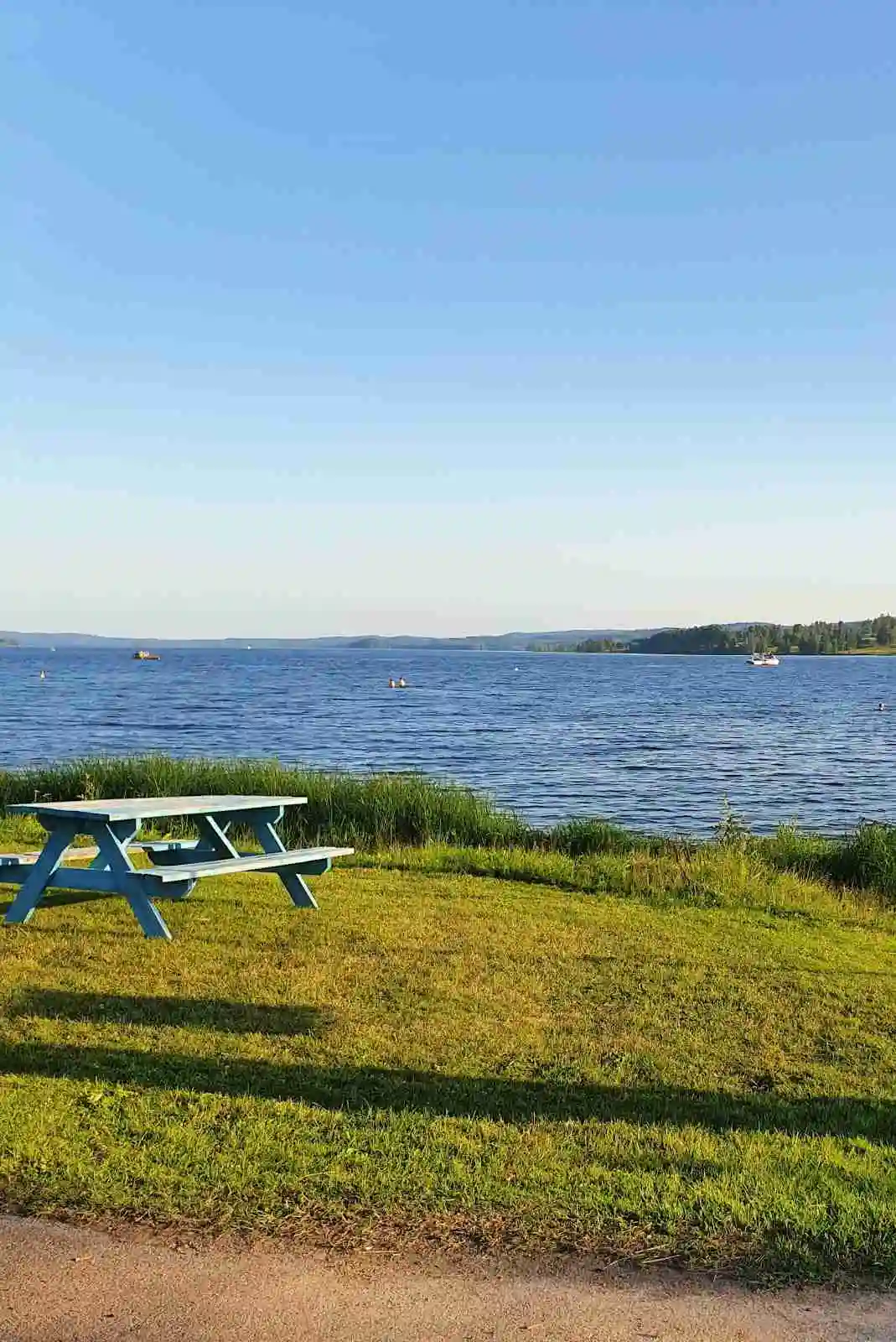 En vy över en blå sjö med en horisont i fjärran. Vid strandkanten finns en utomhusbänk och annan utemöblering, omgiven av kustlinje och naturlandskap.