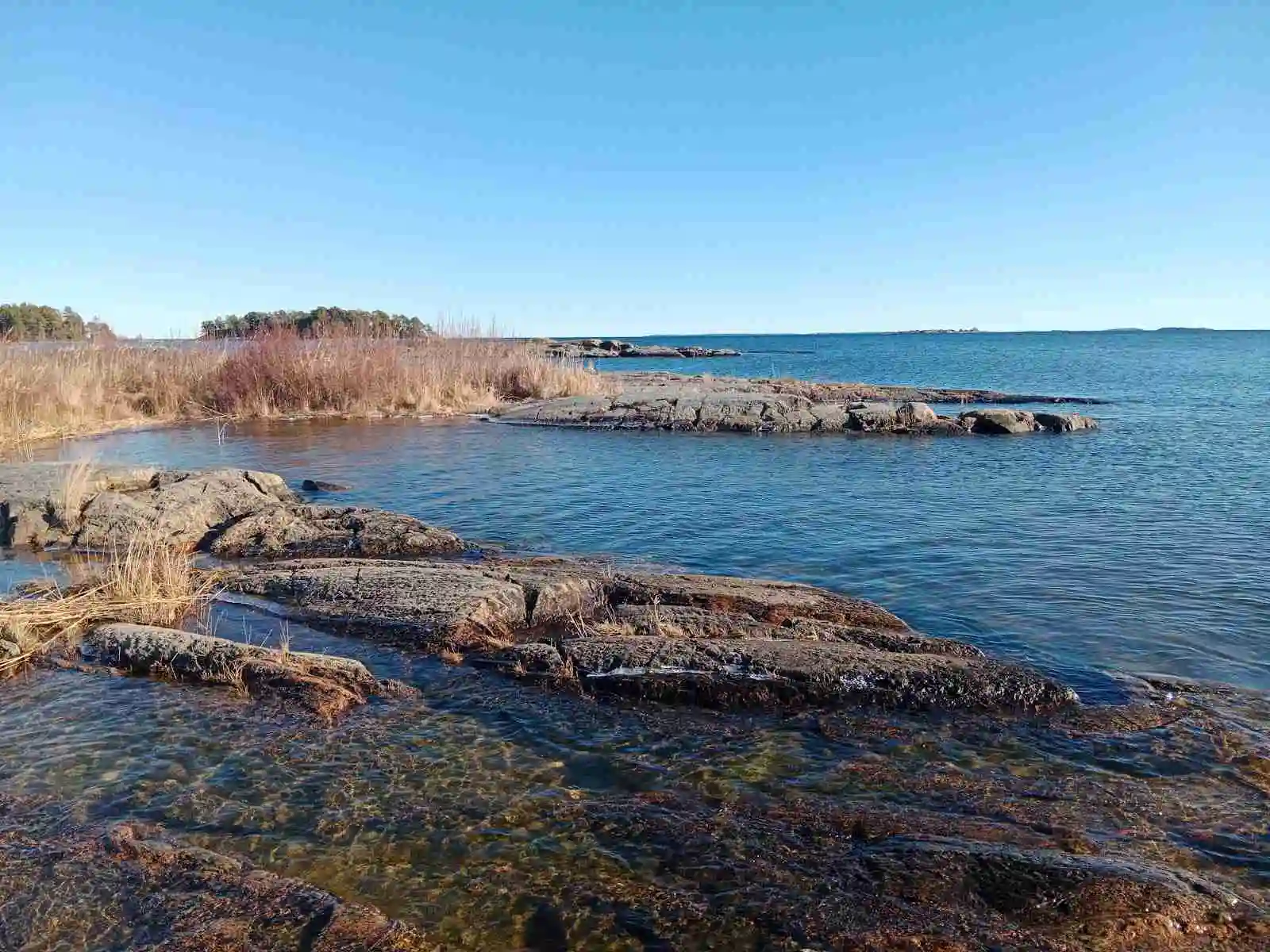 Vy över Ekenäs gästhamn & camping med blått vatten i förgrunden, omgivna av kustlinje och klippor under en klar horisont.