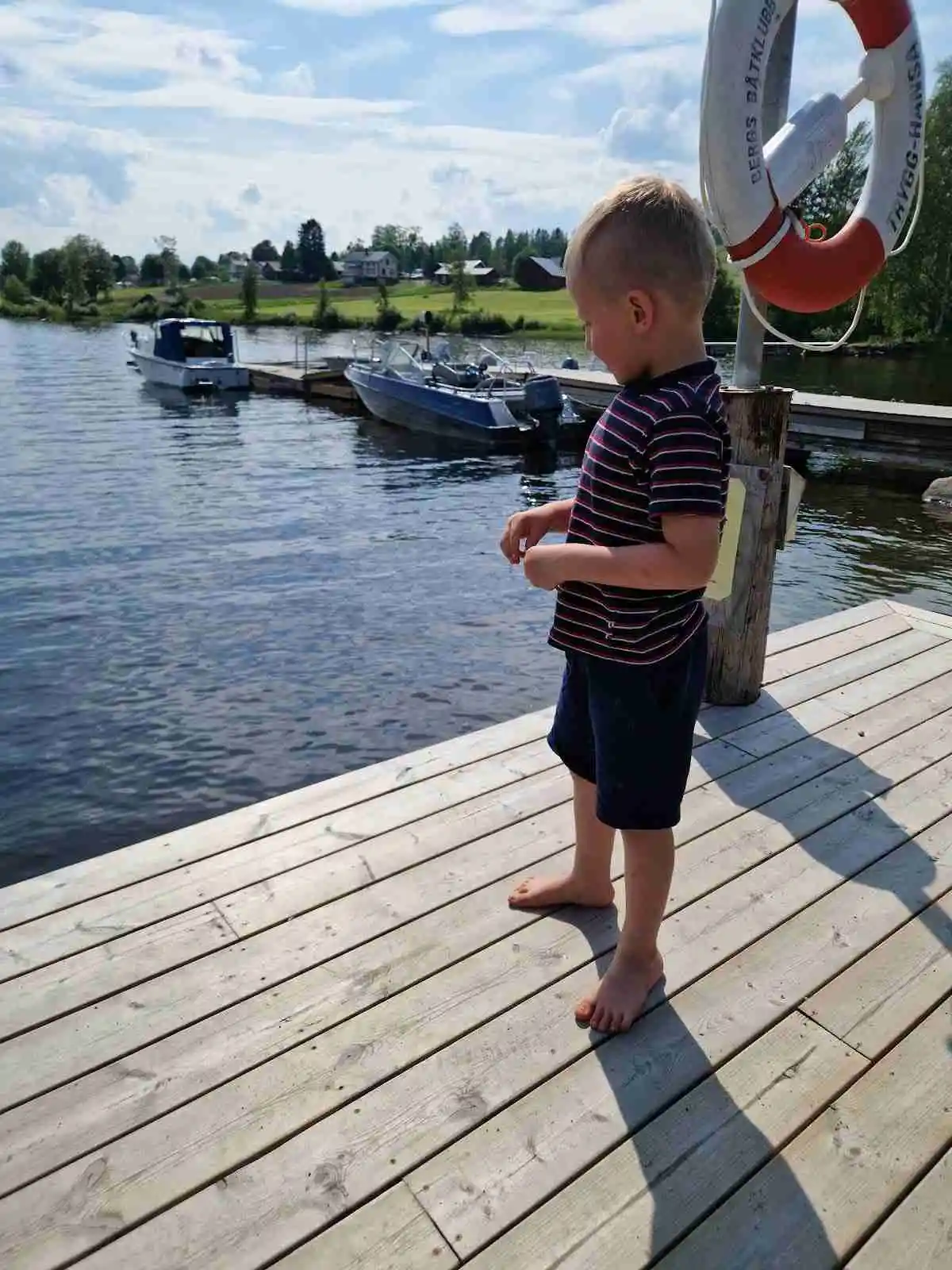 En bild av ett litet barn i shorts som leker vid en brygga en solig sommardag. I bakgrunden syns en liten båt och vattnet glittrar i solskenet, vilket ger en känsla av sommar och semester vid Hoverbergs camping.