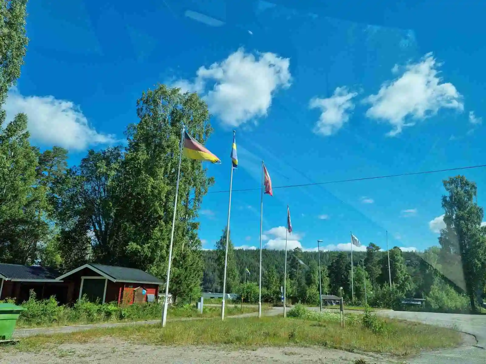 En campingplats med frodig gräsmatta omgiven av träd och buskar. Det finns en flaggstång i närheten och det är vår i luften. I bakgrunden syns tallar som bidrar till den naturnära atmosfären.