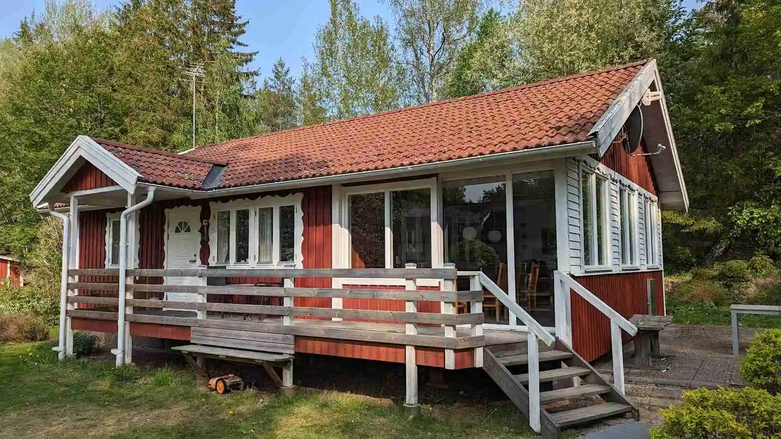 Bild av en mysig timmerstuga omgiven av grönska på Hjorthålan Camping. Stugan har ett tak med verandatak och träfasad, ligger på en välskött tomt med trädgårdsbyggnader i närheten.