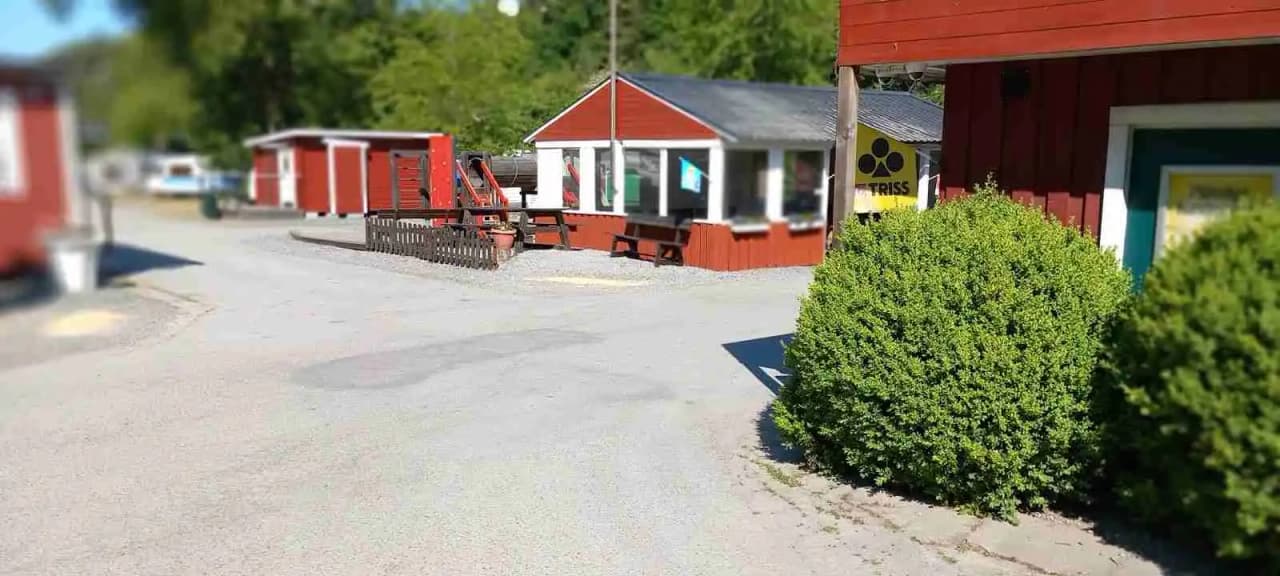 En stuga vid Ängby camping med en gräsmatta och en grusad uppfart, omgiven av träd och sittplatser för avkoppling.