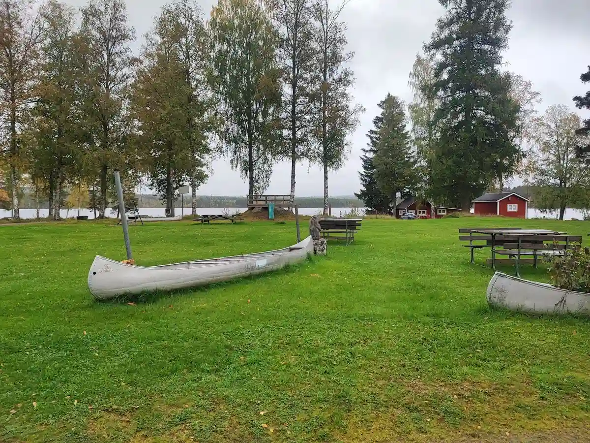 Bild av en grönskande gård i Värmland med en stor gräsmatta och parkmiljö. I förgrunden står en utomhusbänk och i bakgrunden syns en båt och kanot vid vattnet.