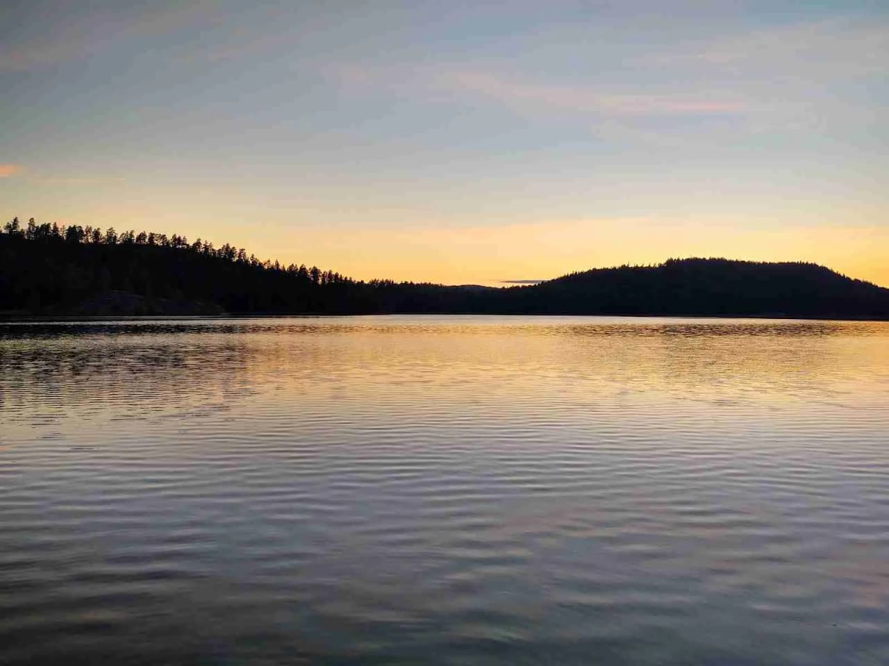En naturskön vy vid Pinnarp Camping med en stilla vattenyta i förgrunden, omgiven av skog under en skymningshimmel.