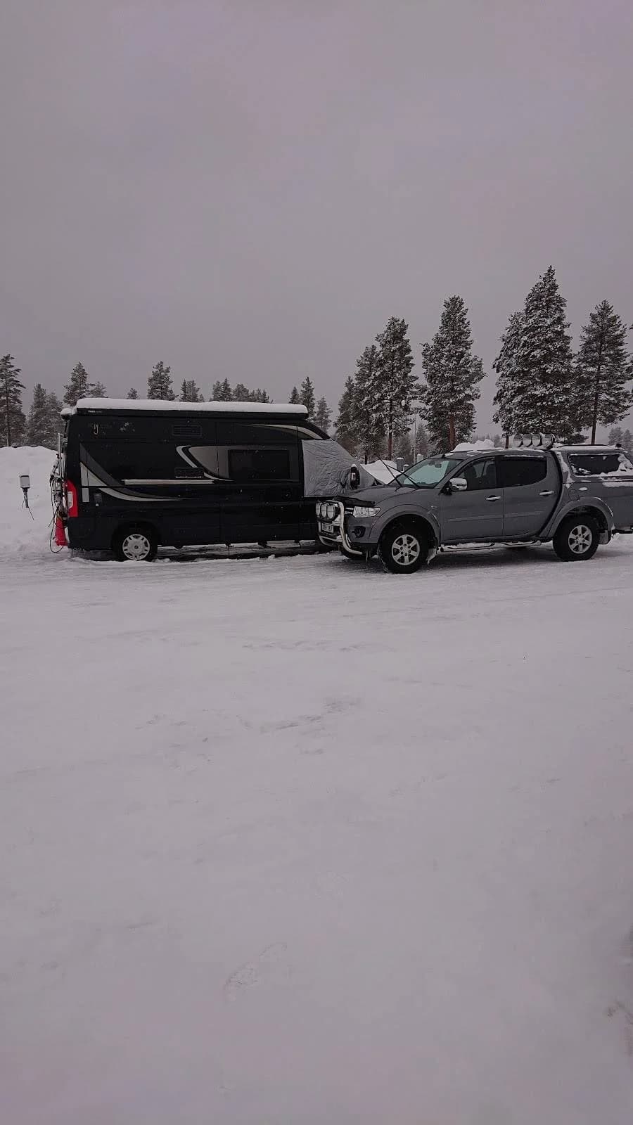 En SUV står parkerad vid en vintrig campingplats, täckt av snö och frost. Träden runtomkring är frostiga, och det kalla vädret skapar en fridfull vinterstämning på Fromhedens camping.