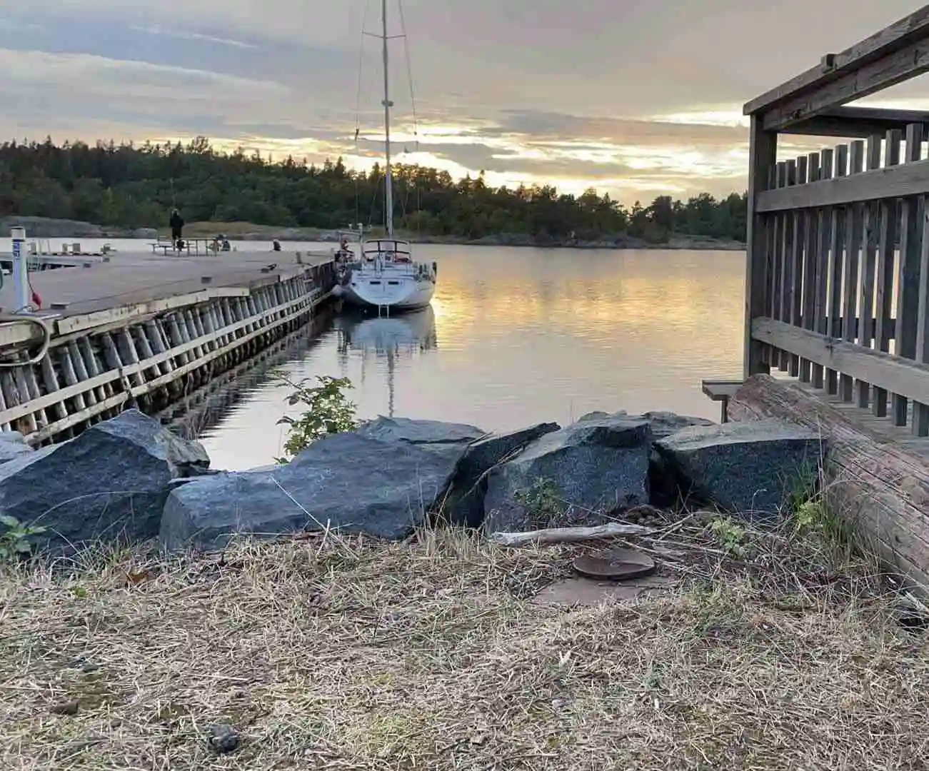 En lugn kustvy vid hamncampingen i Ängskär med flera småbåtar förtöjda vid bryggan i småbåtshamnen, omgiven av glittrande vatten och en bro som syns i bakgrunden.