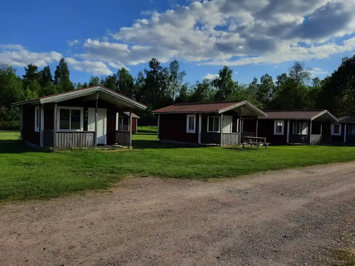 En grön gräsmatta leder fram till en mysig stuga omgiven av träd och buskar på Kosta Bad och Camping. I bakgrunden syns fler byggnader och öppen mark, vilket ger en inbjudande och naturnära känsla på detta besöksmål.