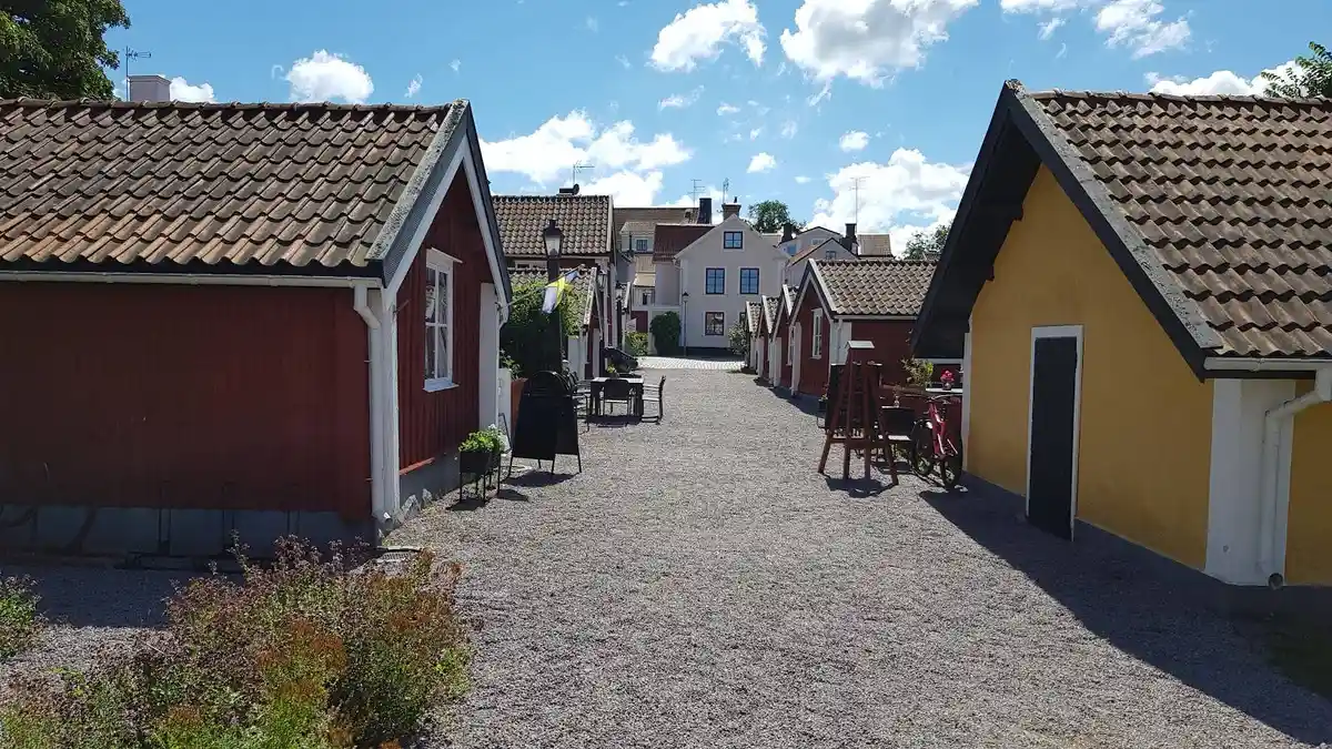 En liten stuga med rödmålad fasad och vita knutar står vid sidan av en asfalterad väg. Stugans tak är belagt med tegel och en dörr syns på framsidan. Framför stugan finns en stenlagd uteplats omgiven av en grönskande gräsmatta.