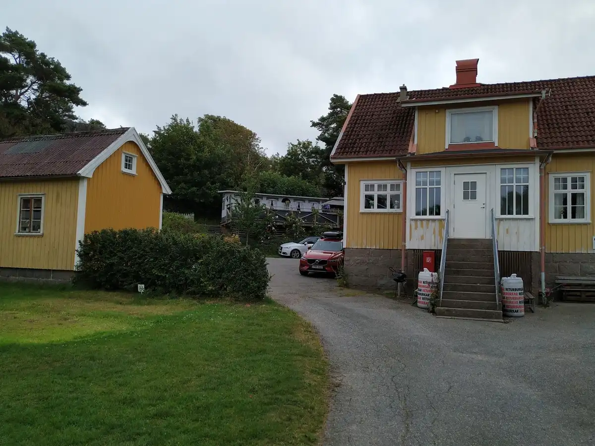 En bild av Löfgrens camping som visar ett rött hus med vitt tak i en lantlig och idyllisk miljö. I bakgrunden syns moln mot en ljus himmel och omgivningen präglas av grönska och andra småbyggnader.