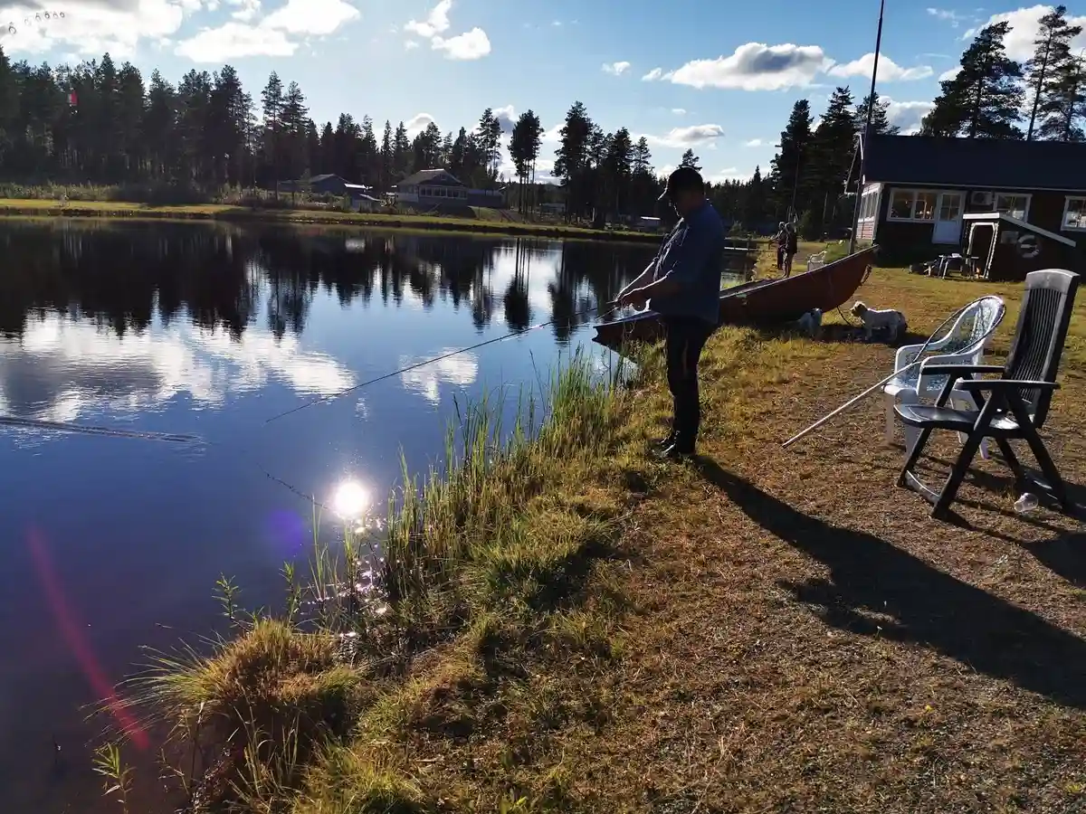 En stilla insjö med spegelblanka vatten omgiven av gröna träd. En ensam fiskare står vid sjöns kant och njuter av den fridfulla omgivningen vid Fromhedens camping.