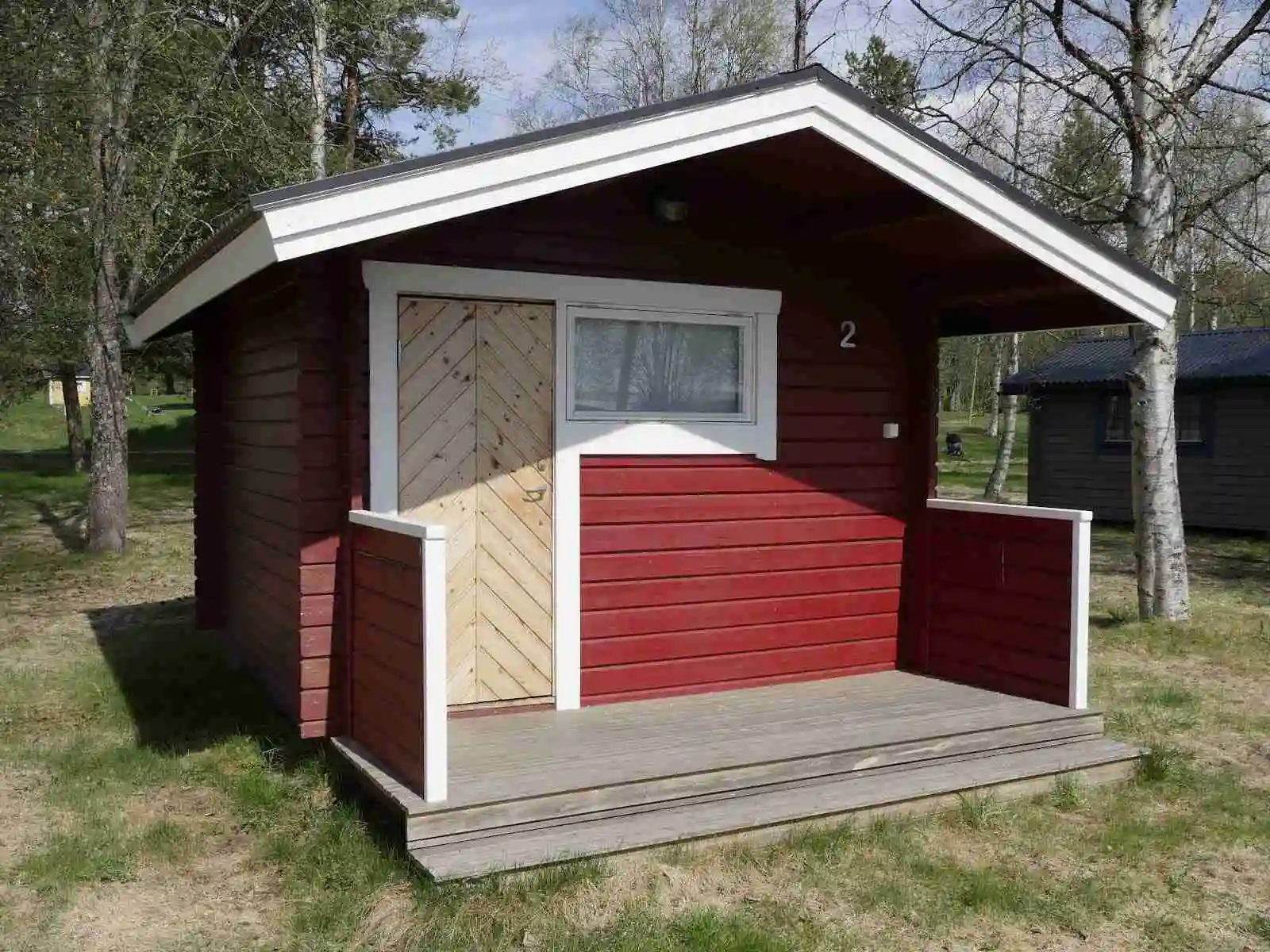 Bild av en trästuga vid Hoverbergs camping. Stugan är byggd med plankor av trä och har en liten veranda. Träets naturliga färger och strukturer är synliga, vilket ger en rustik och inbjudande känsla. Bakgrunden består av omgivande natur som omsluter stugan.