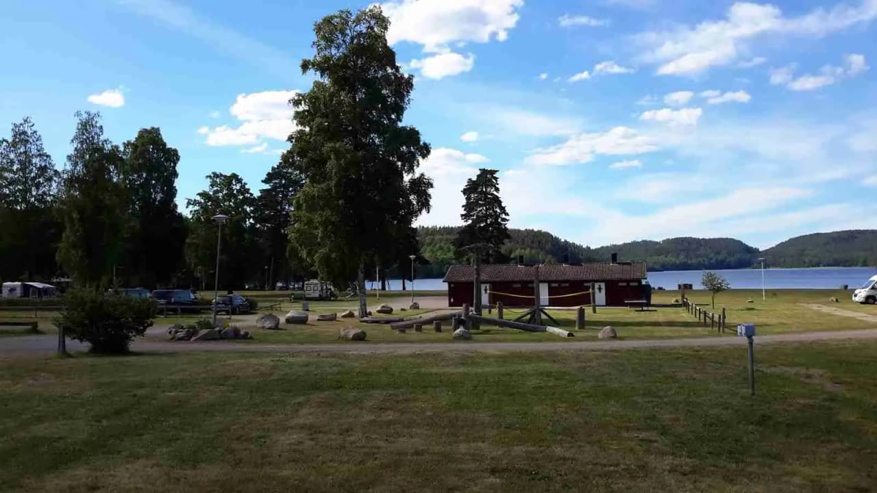 En glänta i en campingmiljö med gröna gräsmattor, en picknickbänk och ett lågt molntäcke över en klarblå himmel.