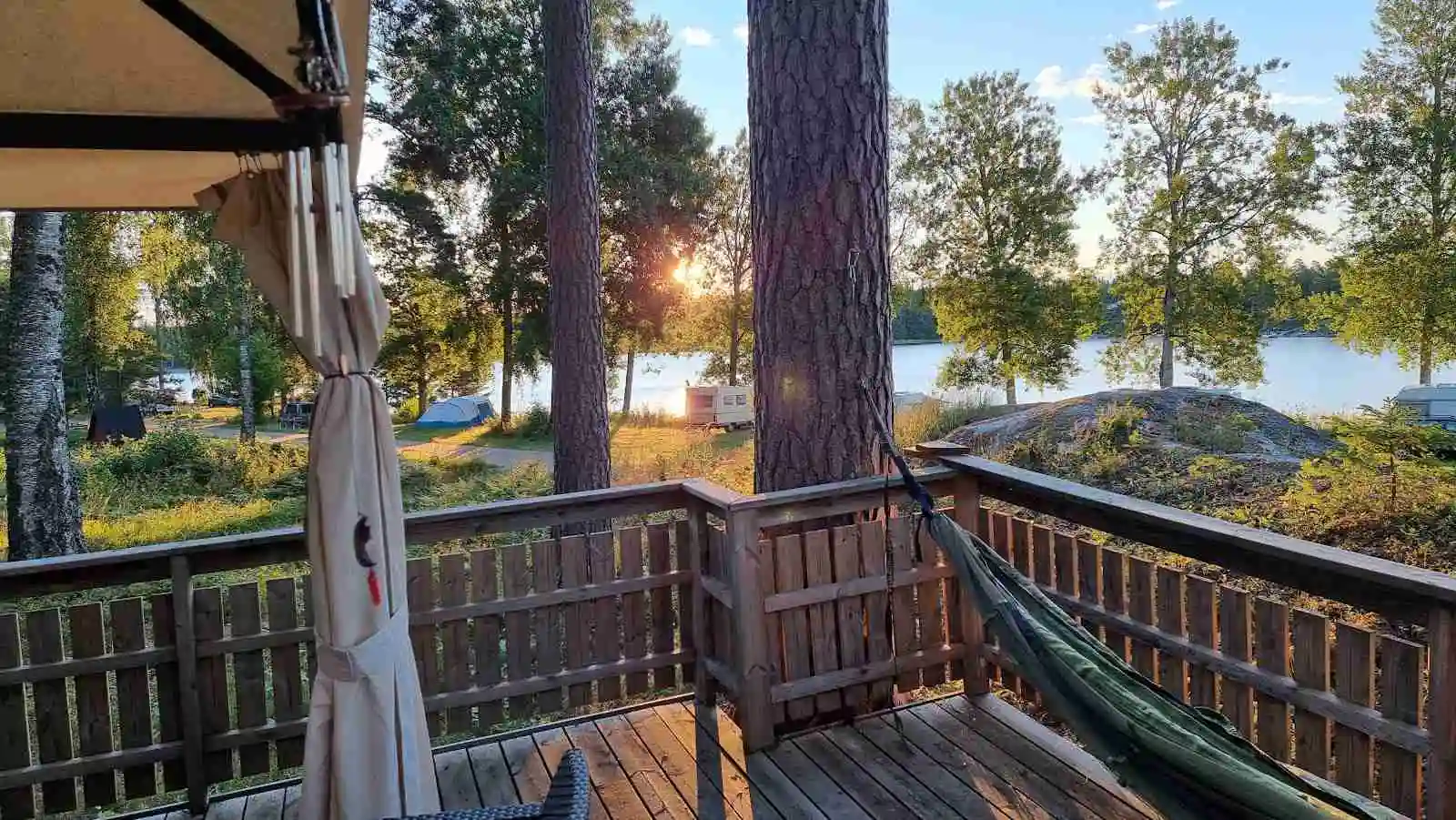 En trästuga med en veranda omgiven av skuggiga träd, belägen vid ett vattenreservoar. På verandan finns stolar som inbjuder till avkoppling, omgiven av natur. Perfekt för en ekologisk campingupplevelse på Elovsbyn Camping & Canoe.