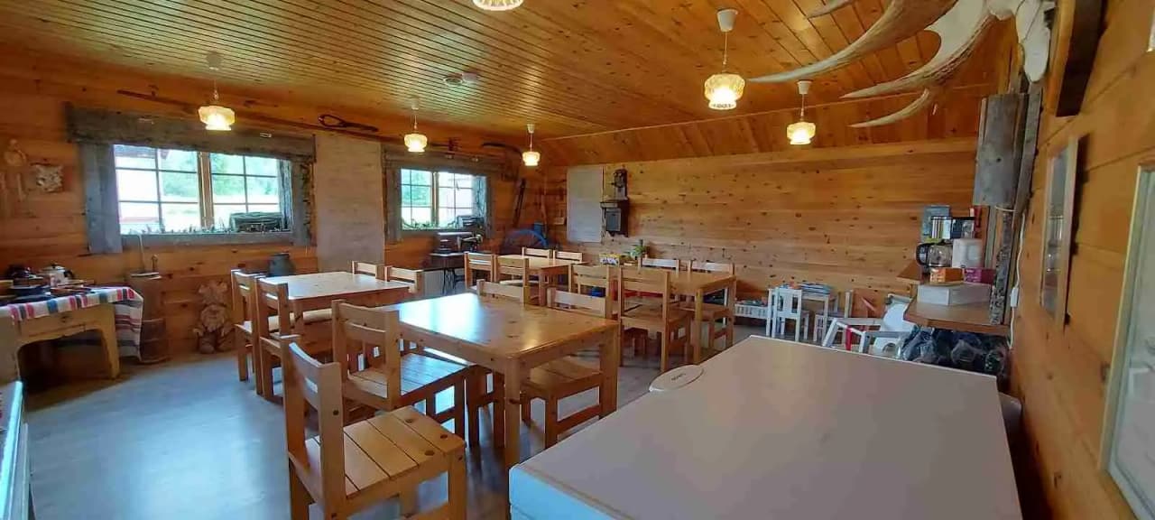 En mysig interiör av en trästuga med synliga bjälkar och träpaneler. I förgrunden finns ett köks- och matrum med trämöbler, inklusive ett matbord och stolar. Miljön påminner om en log cabin-stil, vilket ger en rustik och hemtrevlig atmosfär.