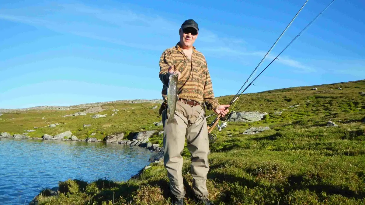 En person står vid kanten av en sjö på Rörvattnets fiskecamp och kastar sin fiskelina med ett metspö i det stilla vattnet, omgiven av vacker natur.